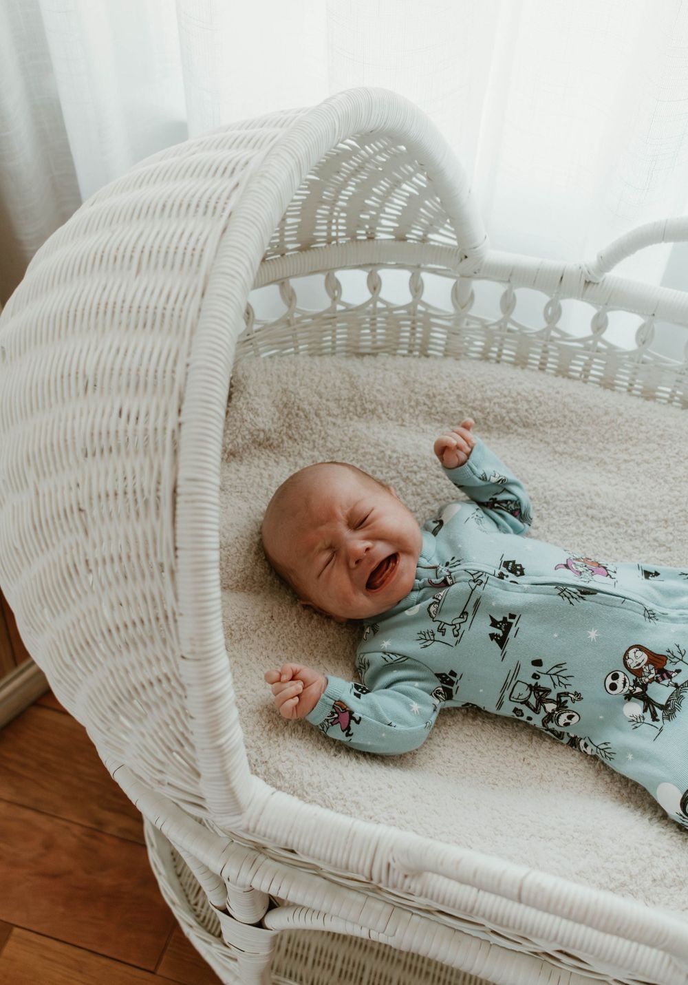 Baby wearing blue patterned sleeper lying in white wicker bassinet while crying.