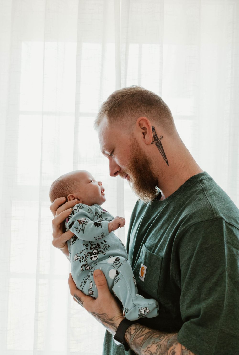 Person in green shirt holding small infant wearing light blue onesie by bright window.