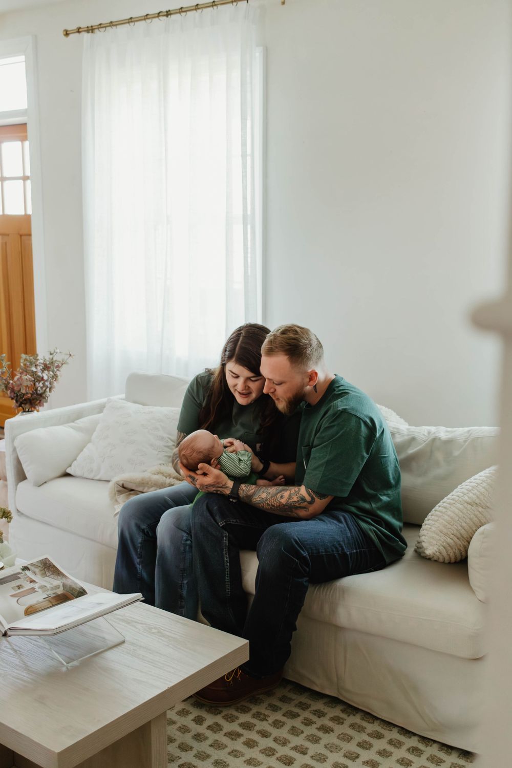 Family shares tender moment with newborn while sitting on white couch in bright living room.