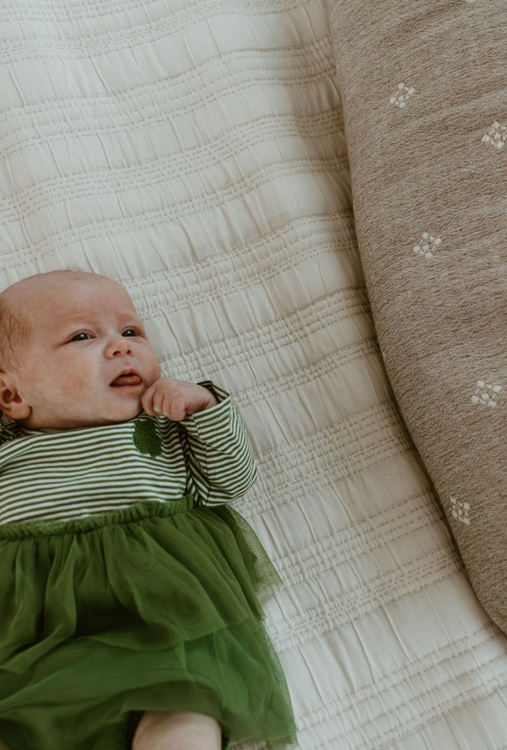 Infant in green dress smiles while lying on white textured bedding.