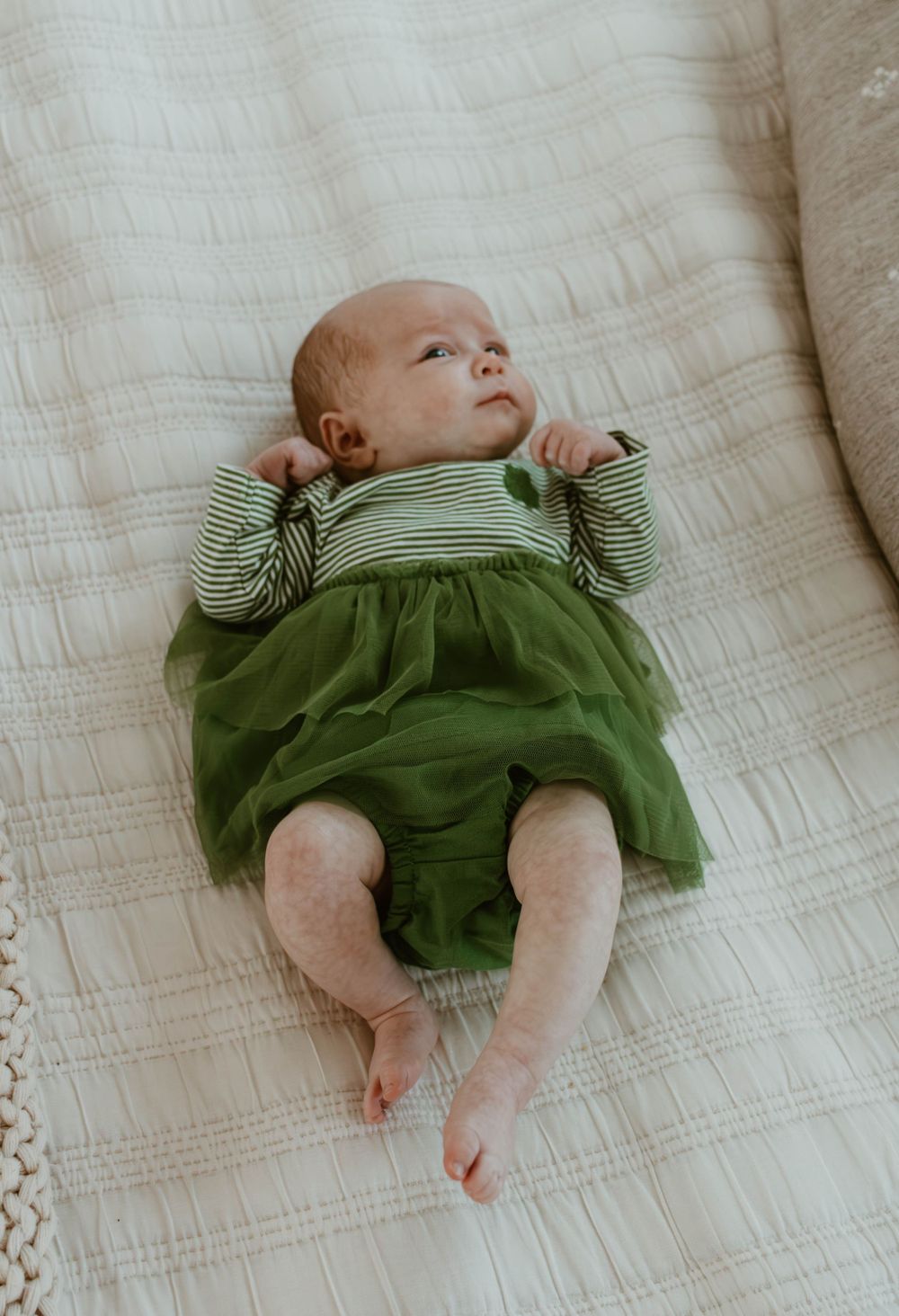 Baby wearing green striped dress gazes upward while resting on white bedding.