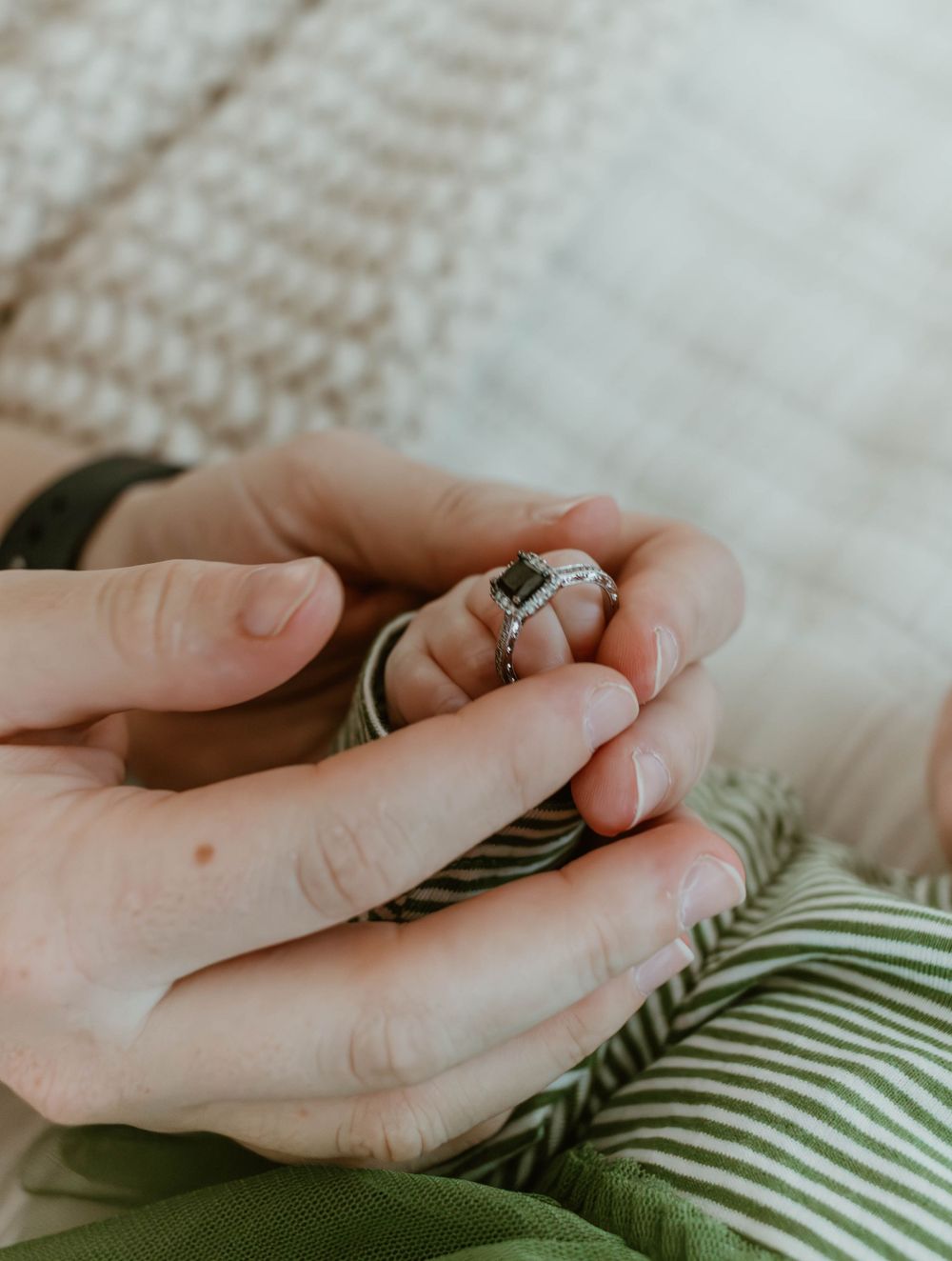 Close-up of hands holding tiny infant fingers with wedding rings visible.