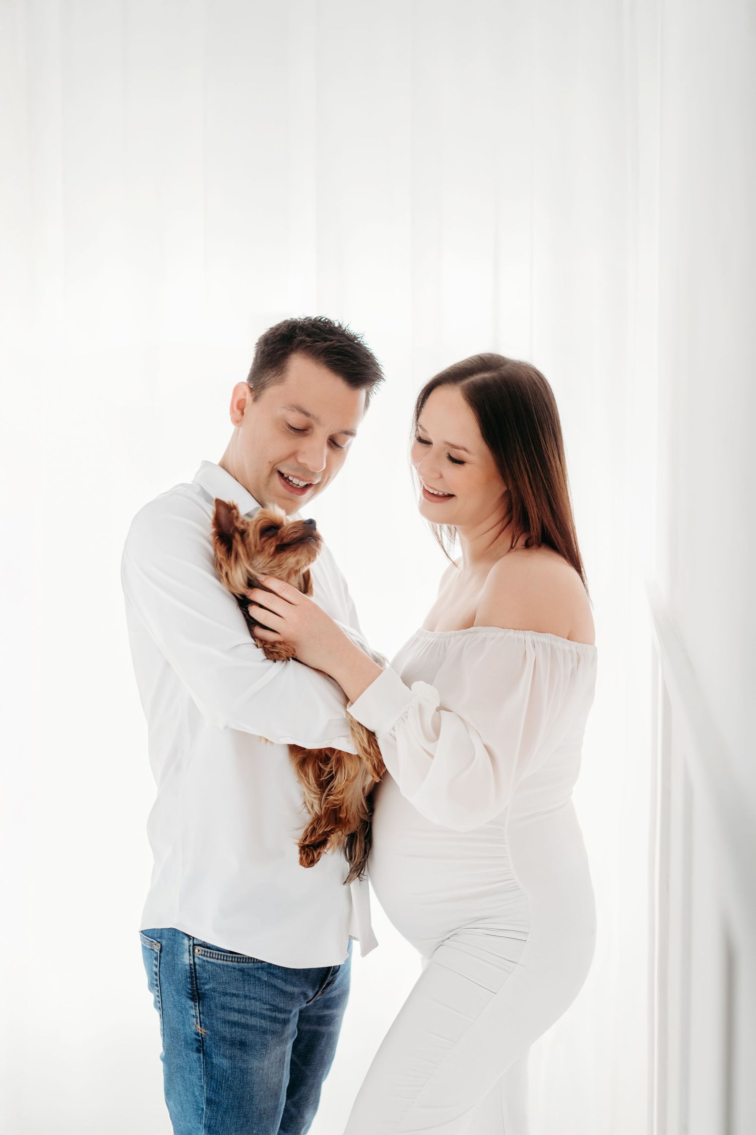 Couple in white outfits holding small pet dog together against light background.