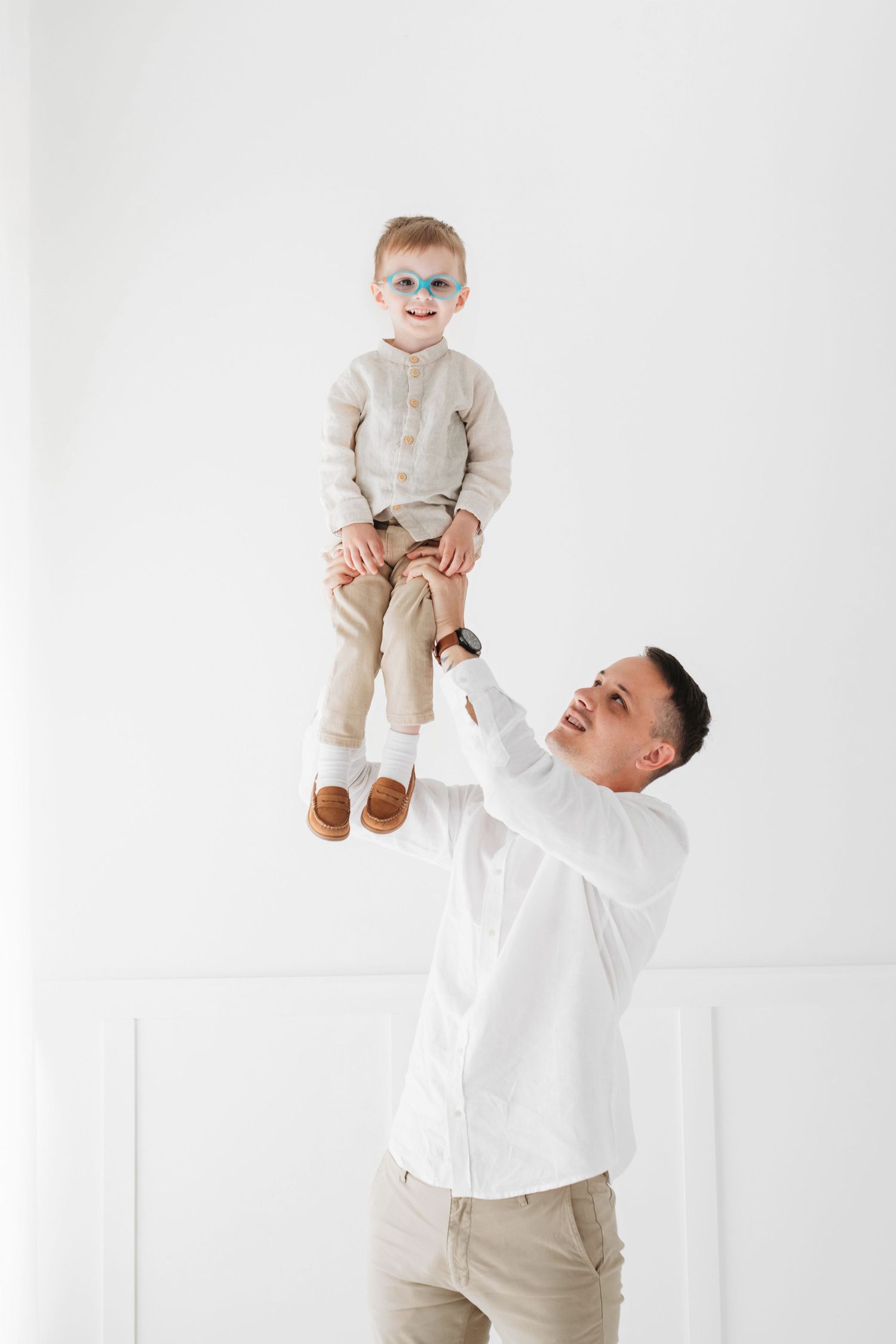 A person in a white sweater lifts a small child wearing beige clothing against a light background.