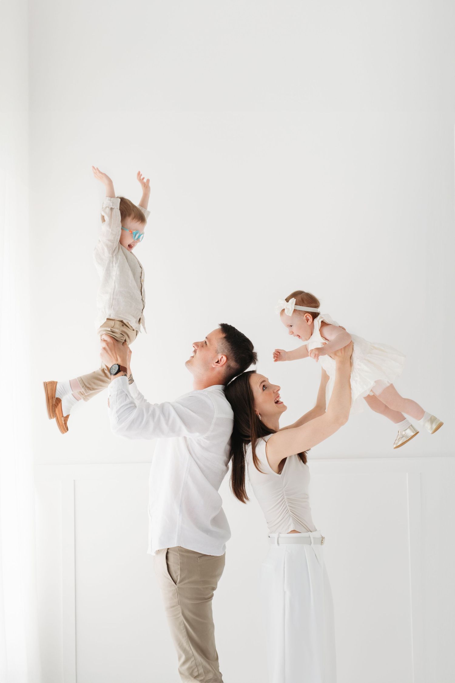 A couple in white shirts and khakis playfully lifting their children against a light background.