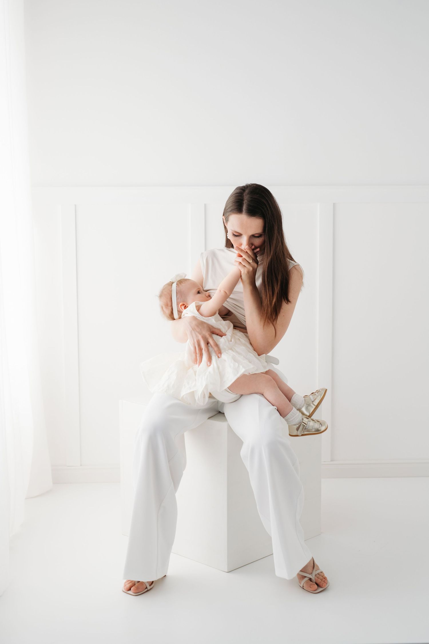 Woman in white outfit sitting on cube holding and interacting with baby in bright minimalist setting.