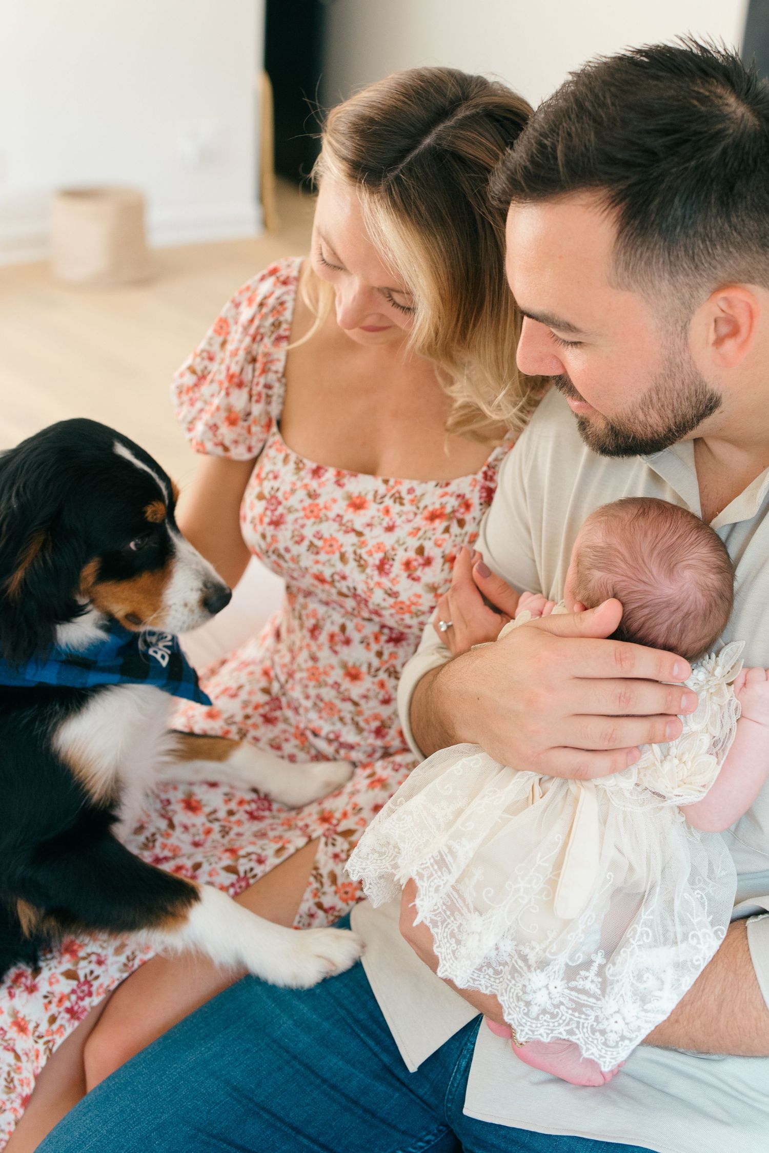 Family of three sitting on bed, holding newborn wrapped in swaddle blanket while the family dog joins.