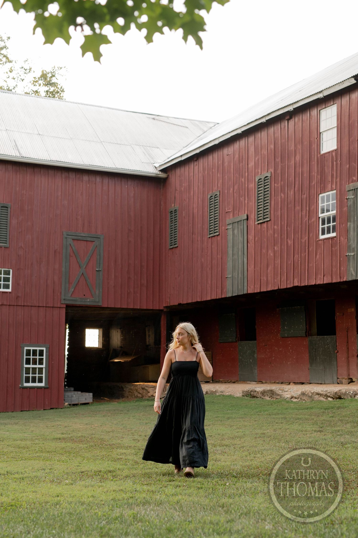 A woman in a long black dress stands in front of a classic red barn on a summer day.