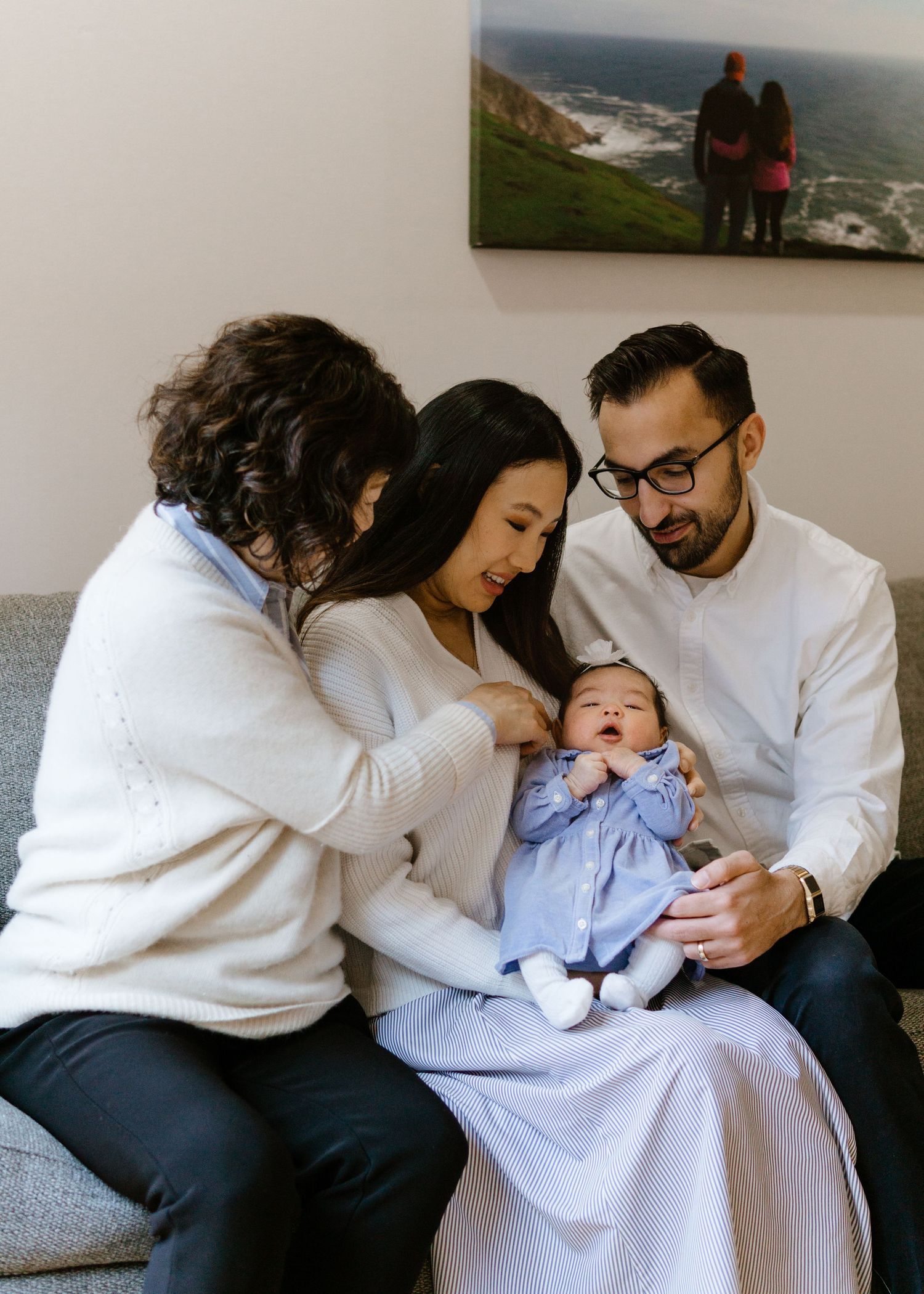 Family gathers on grey couch looking lovingly at their newborn baby dressed in a blue dress.