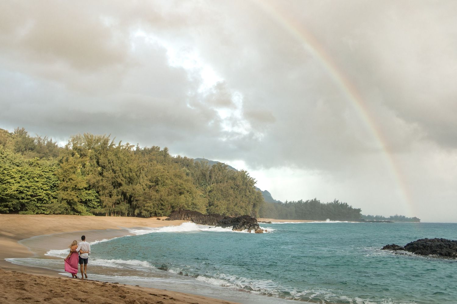 Couple with arms wrapped around one another walking down long white sand beach on Maui with rainbow in the distance