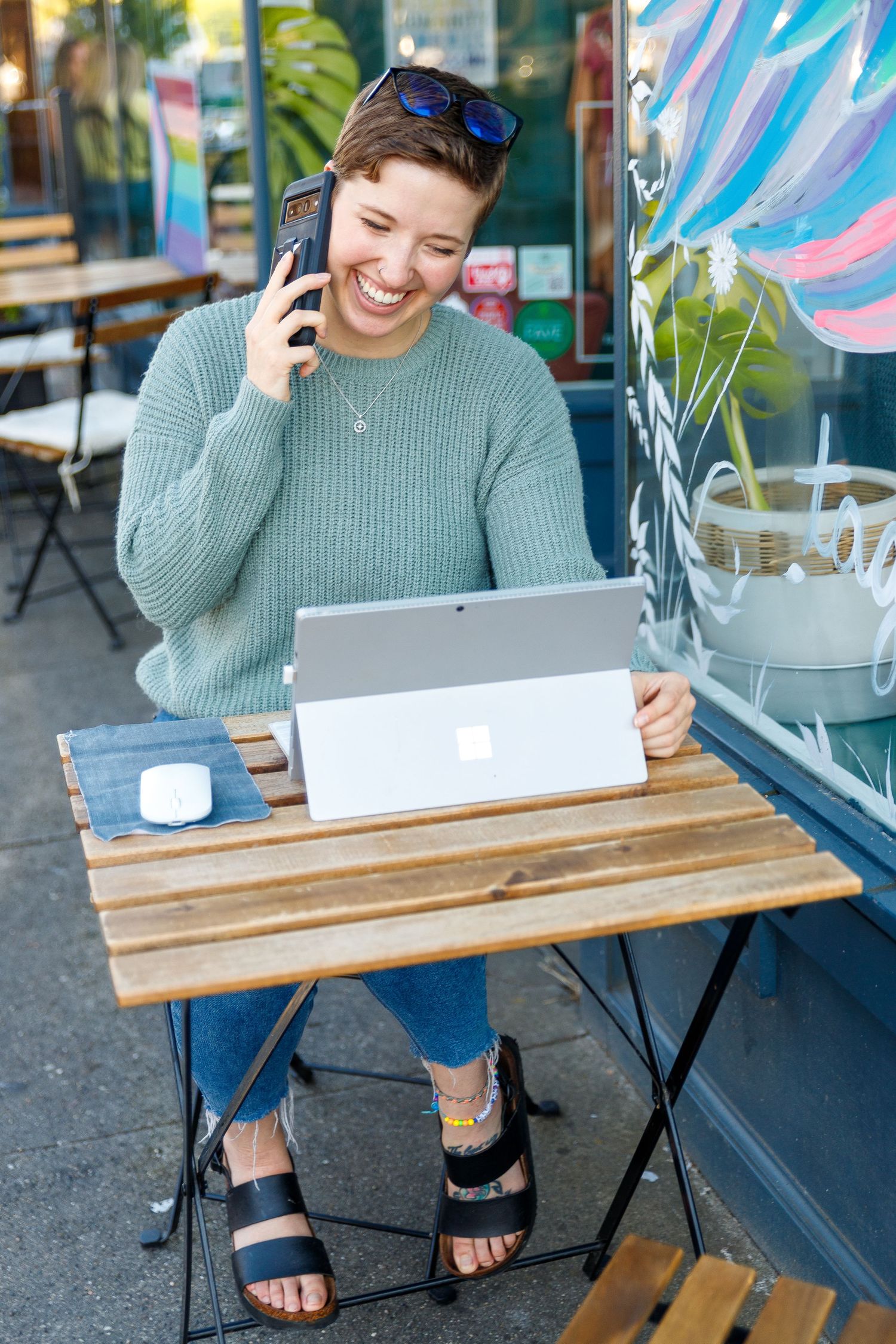 Someone in a gray sweater sits at an outdoor cafe table working on a tablet while talking on the phone.