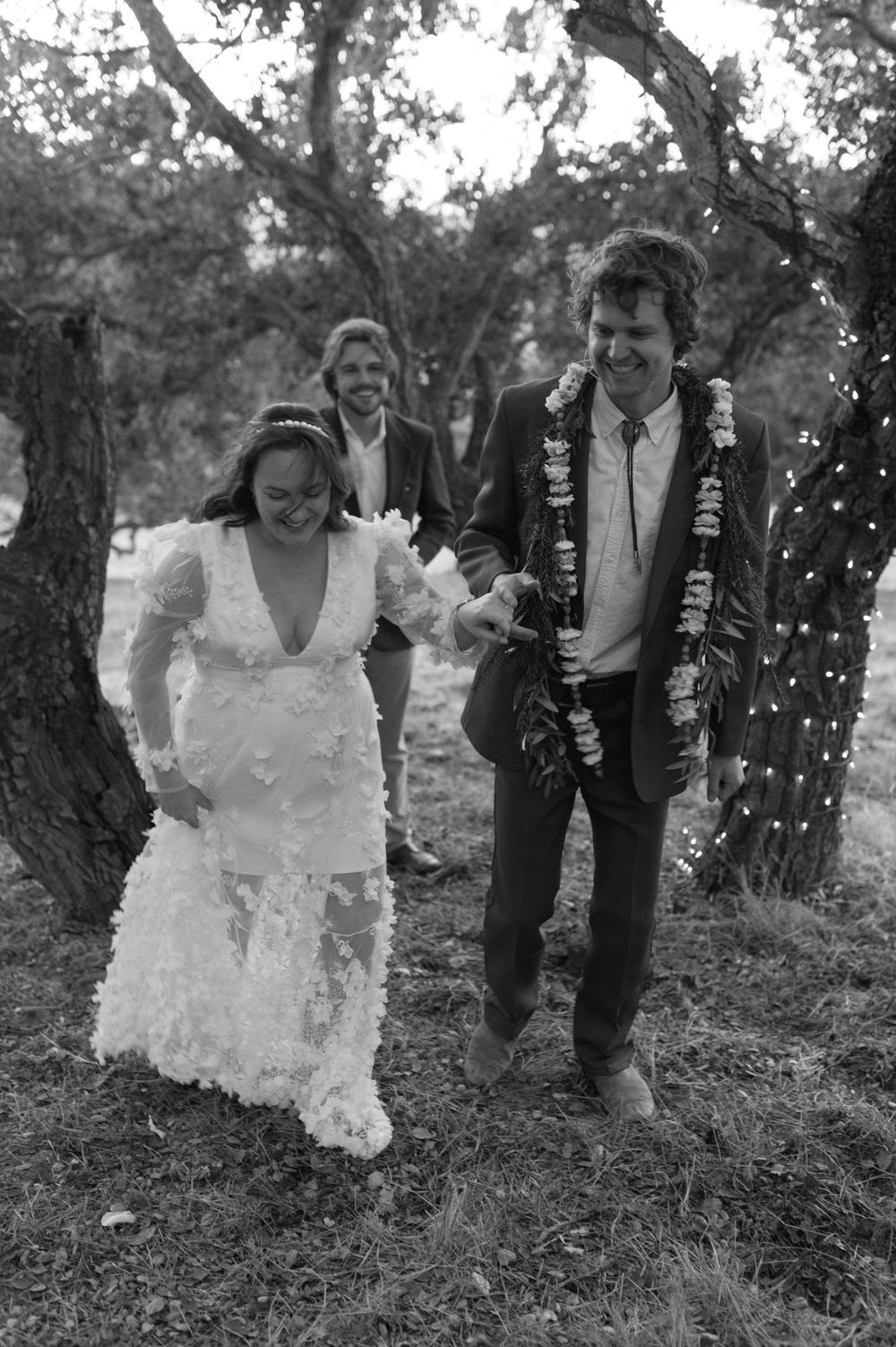 A candid black and white wedding photo showing a couple walking together outdoors with string lights and trees in background.