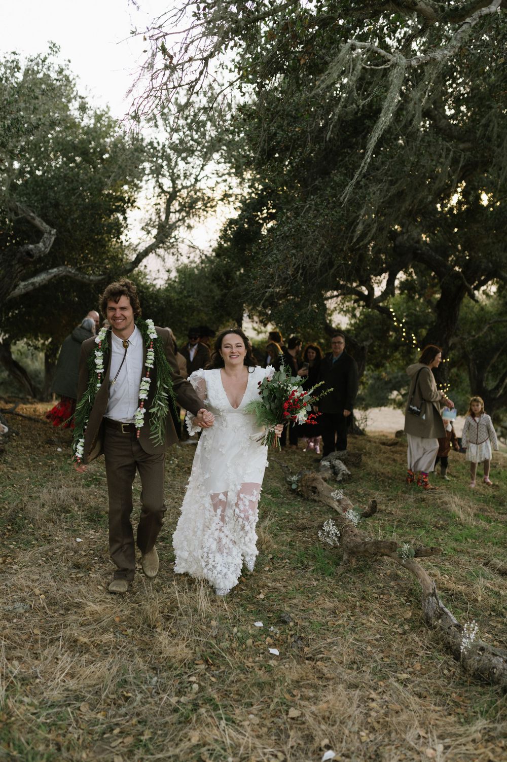 Newlyweds walking down a hillside path surrounded by trees and wedding guests.