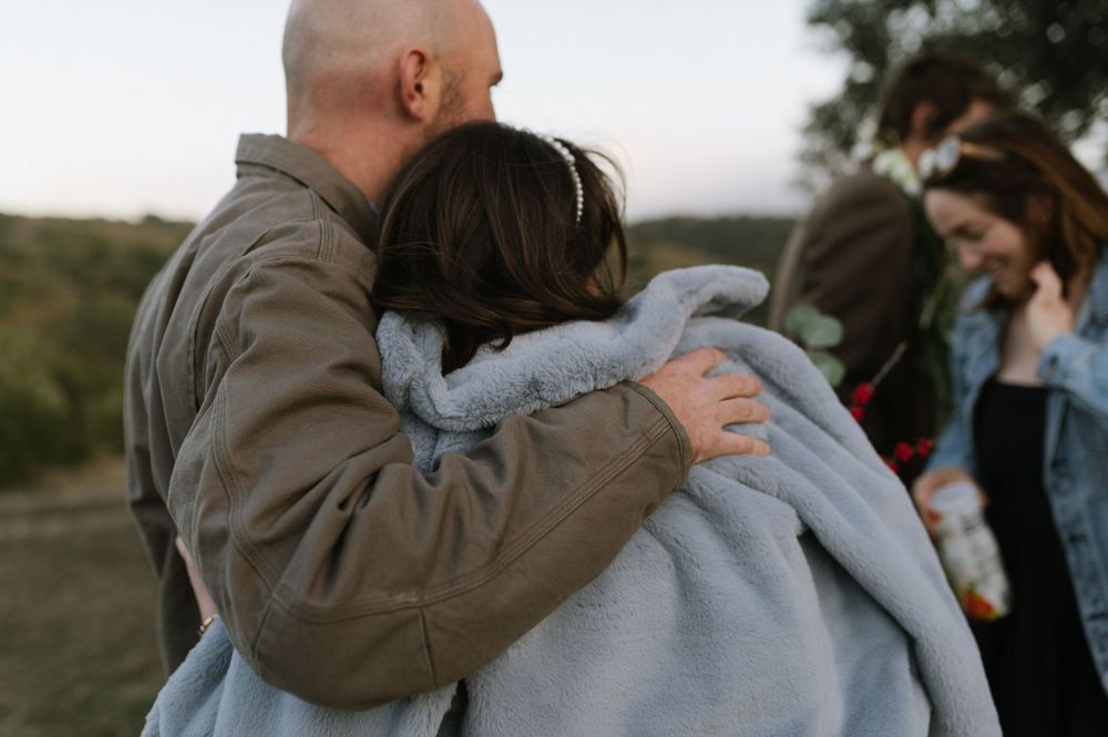 A couple embraces while standing outdoors during sunset with someone visible in the background.