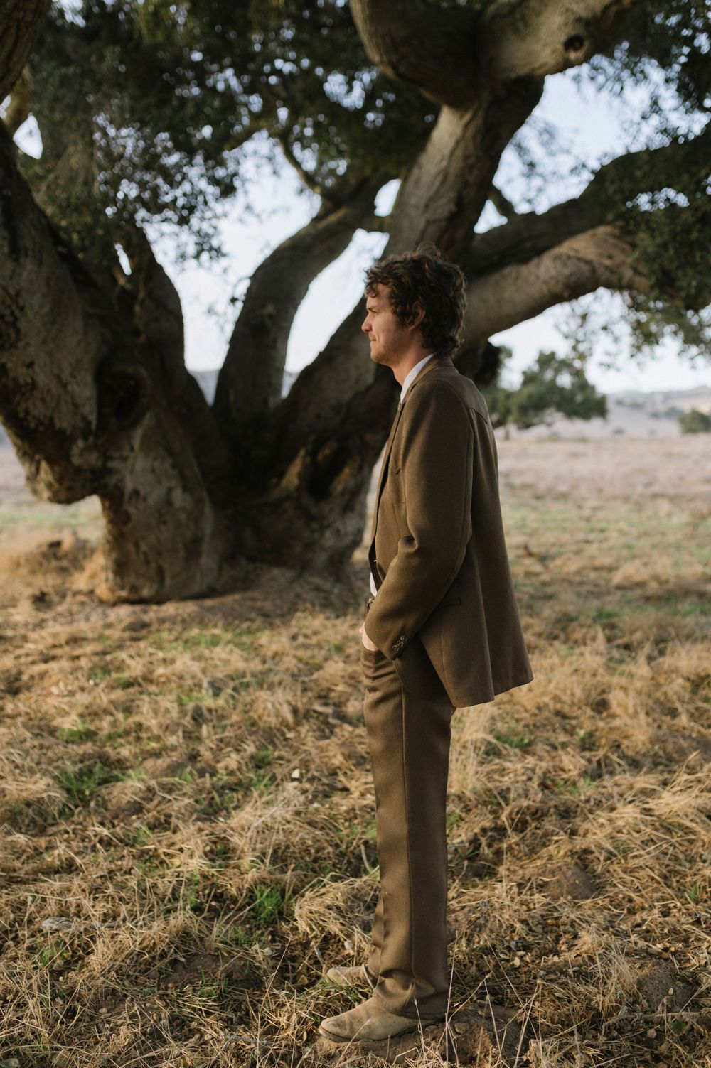 A person in a tan suit stands beneath a large gnarled tree in a golden field.