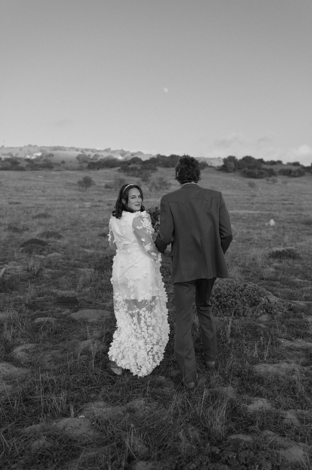 Black and white photo of couple standing in rural field with mountains in background.