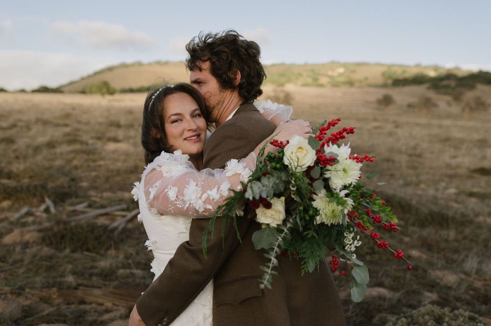 Candid wedding portrait with couple embracing while holding floral bouquet in desert setting.