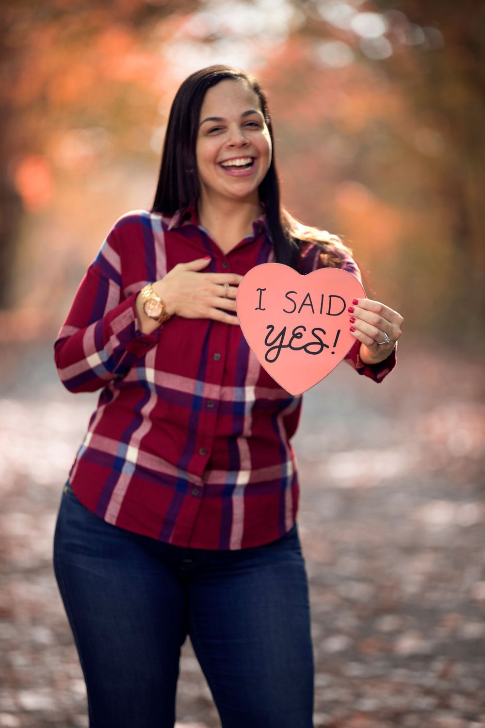 Someone in a plaid red shirt and jeans holds up a pink heart sign with 'I SAID: YES!' during an autumn engagement shoot.