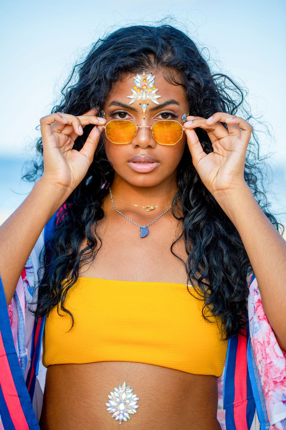 Fashion portrait with yellow sunglasses, orange crop top, ornate white face jewels and floral kimono against blue sky background.