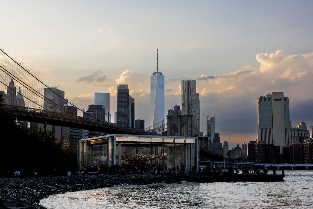 The New York City skyline at sunset featuring One World Trade Center and the Manhattan Bridge over the East River.