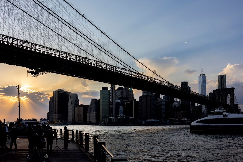 Silhouette of Brooklyn Bridge against Manhattan skyline at sunset with One World Trade Center visible in background.