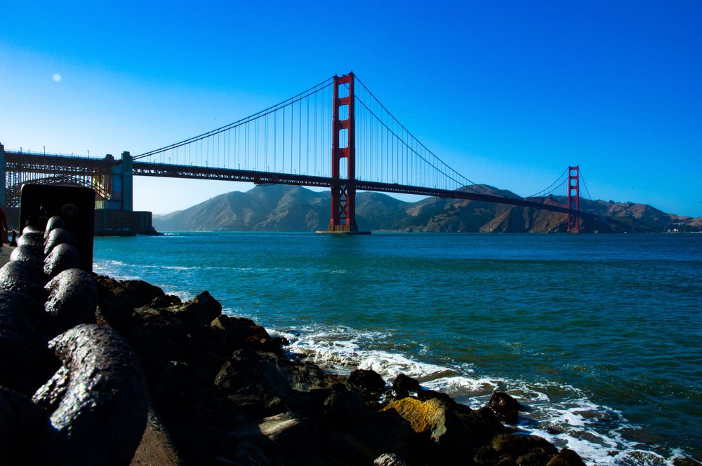 The iconic Golden Gate Bridge spans the San Francisco Bay with turquoise waters and rocky shoreline in the foreground.