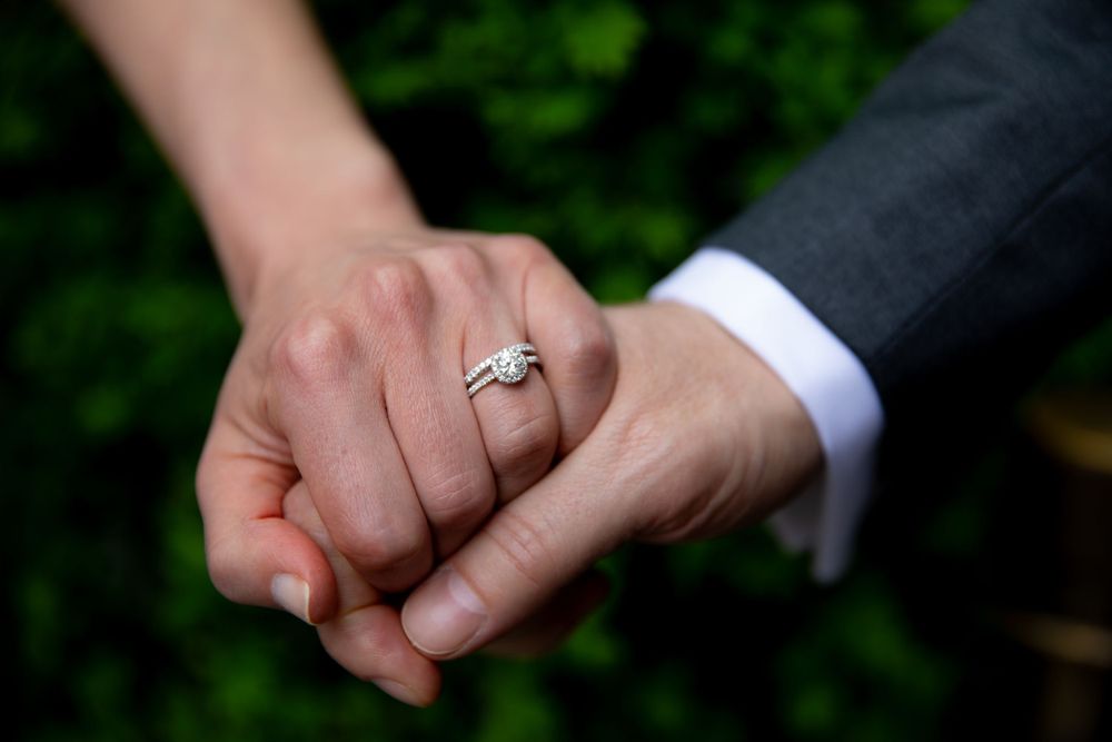 Close-up shot of newlywed couple holding hands with engagement ring against green foliage background.