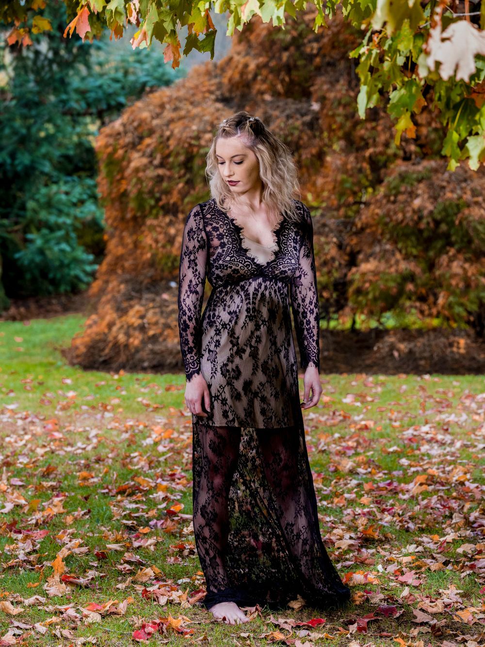 A model poses in a black lace maternity gown on autumn leaves with orange foliage in the background.