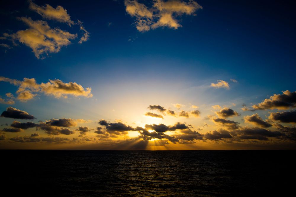 Golden sunset over dark ocean water with dramatic clouds and sunbeams breaking through the evening sky.