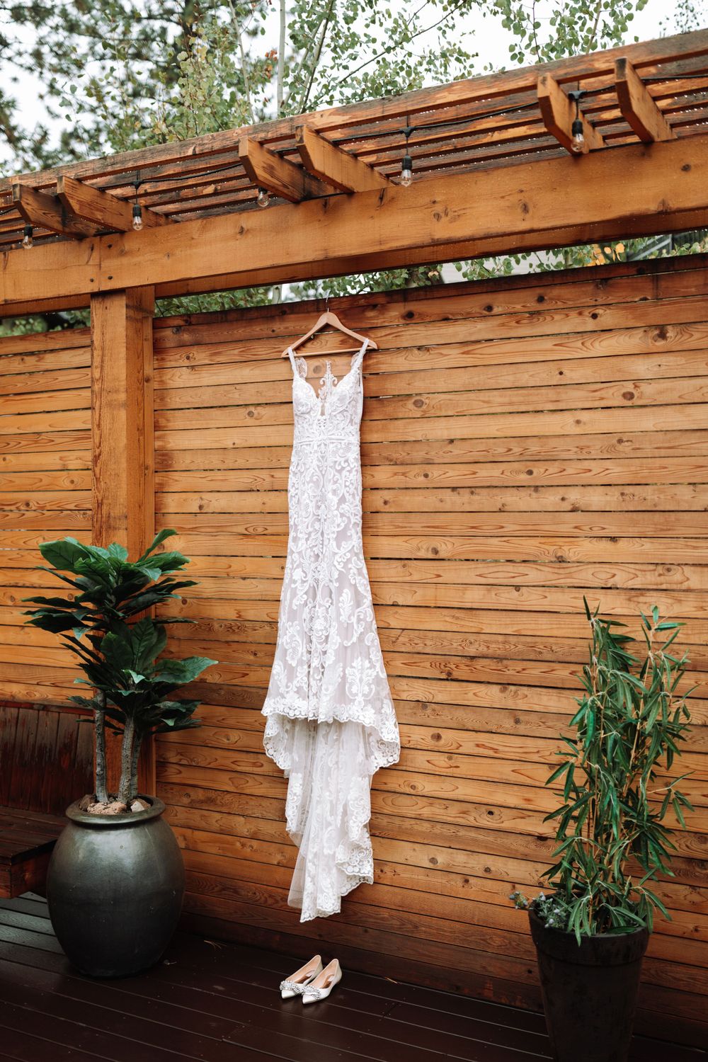 White lace wedding dress hanging from wooden pergola with potted plants on outdoor deck.