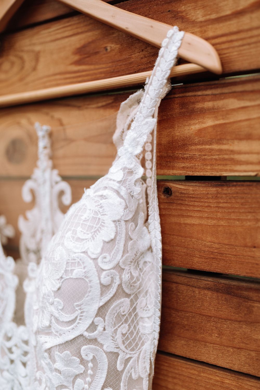 Close-up detail of delicate white lace wedding dress fabric hanging on a wooden hanger against rustic wooden wall.
