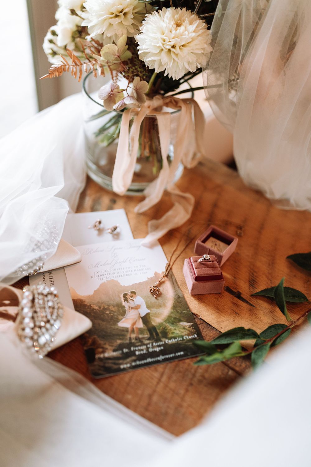 Wedding details including white carnations in a vase, jewelry, rings in a pink box, and tulle on a wooden surface.