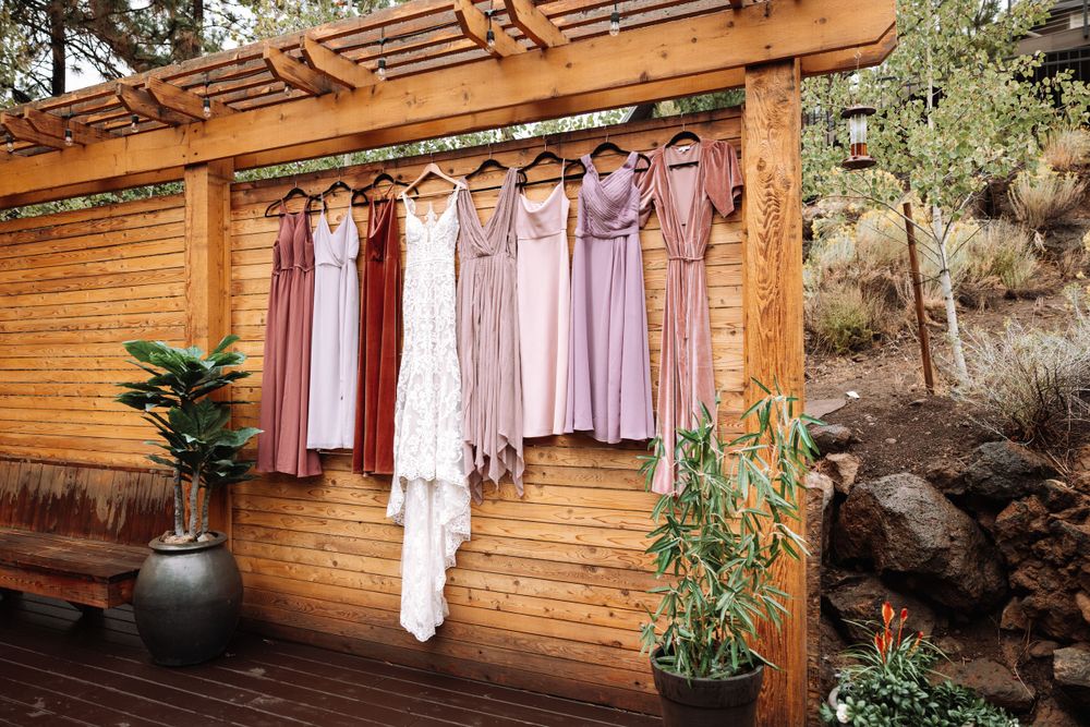 Wedding dresses and bridesmaid gowns in shades of pink and lilac hang on a wooden pergola beside potted plants.