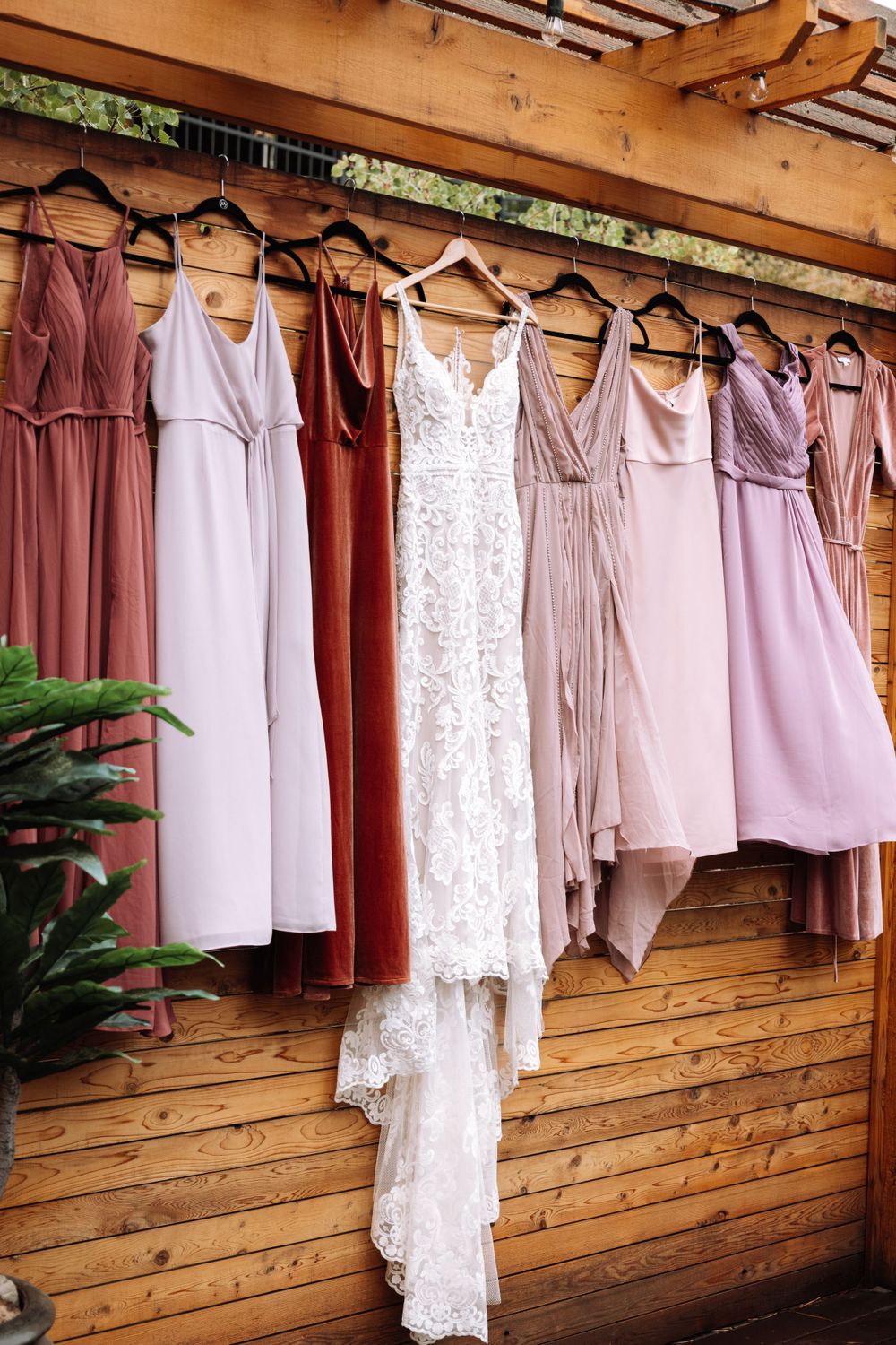 Wedding party dresses in shades of mauve, pink and white with a lace wedding gown hanging on wooden wall.