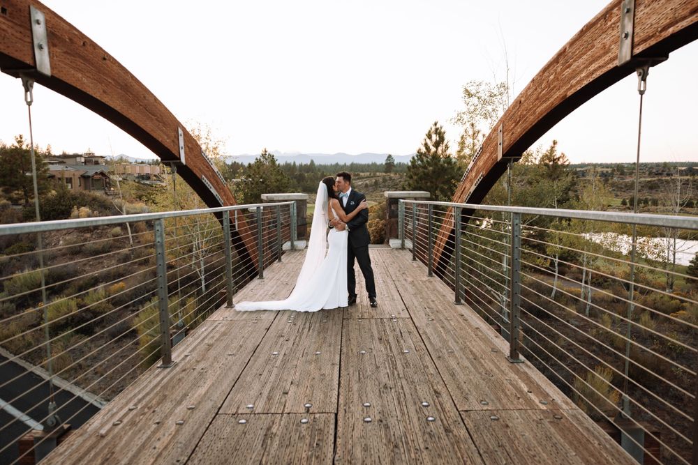A couple stands together on a rustic wooden bridge with curved metal arches overhead against a scenic backdrop.
