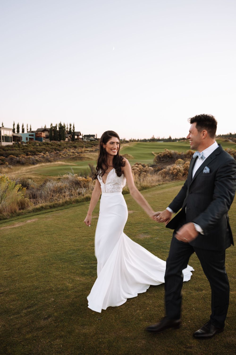 A bride in a flowing white dress dances with her groom on a scenic golf course at sunset.