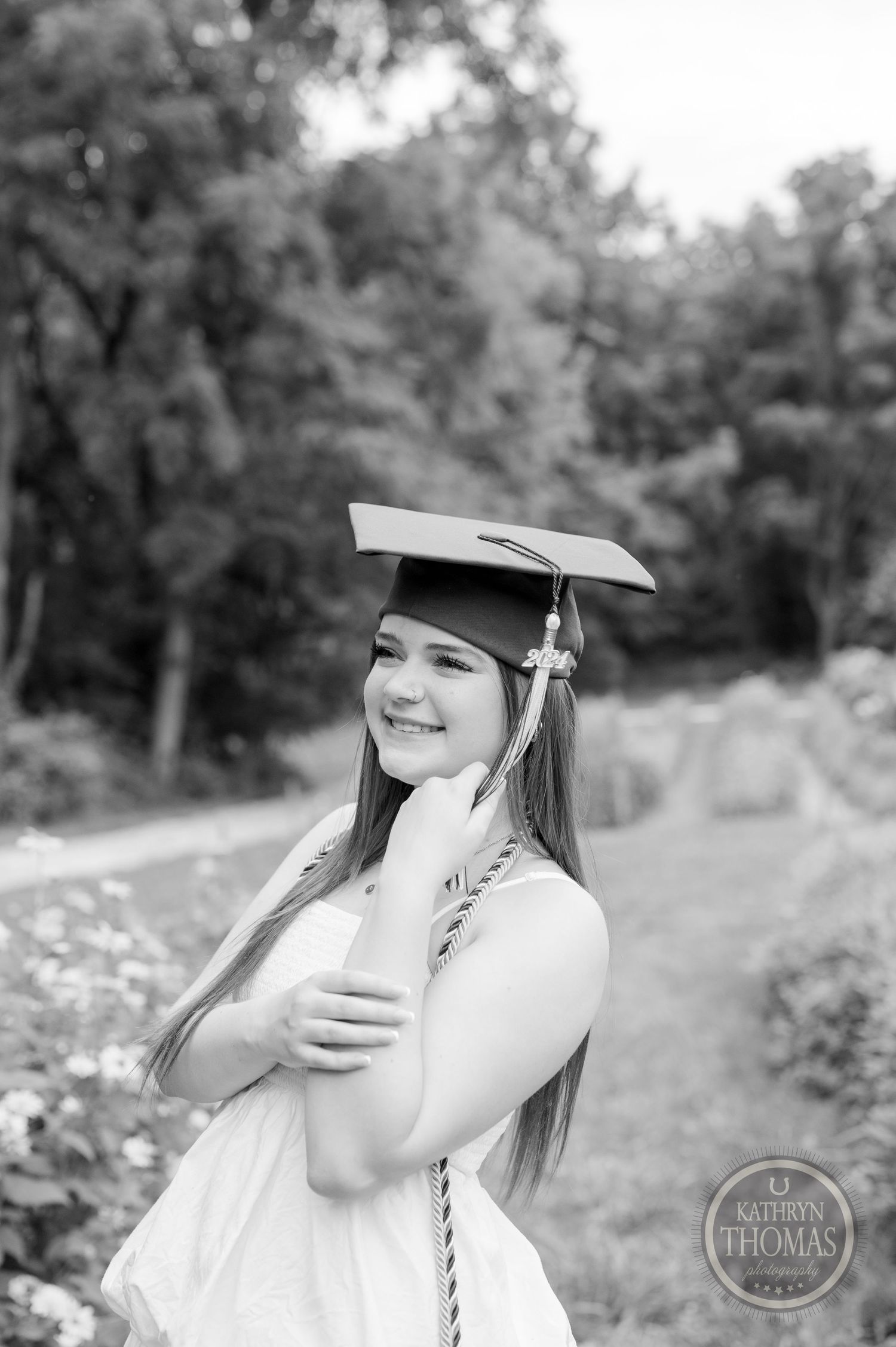 Graduate in white dress and graduation cap poses for portrait in outdoor garden setting.