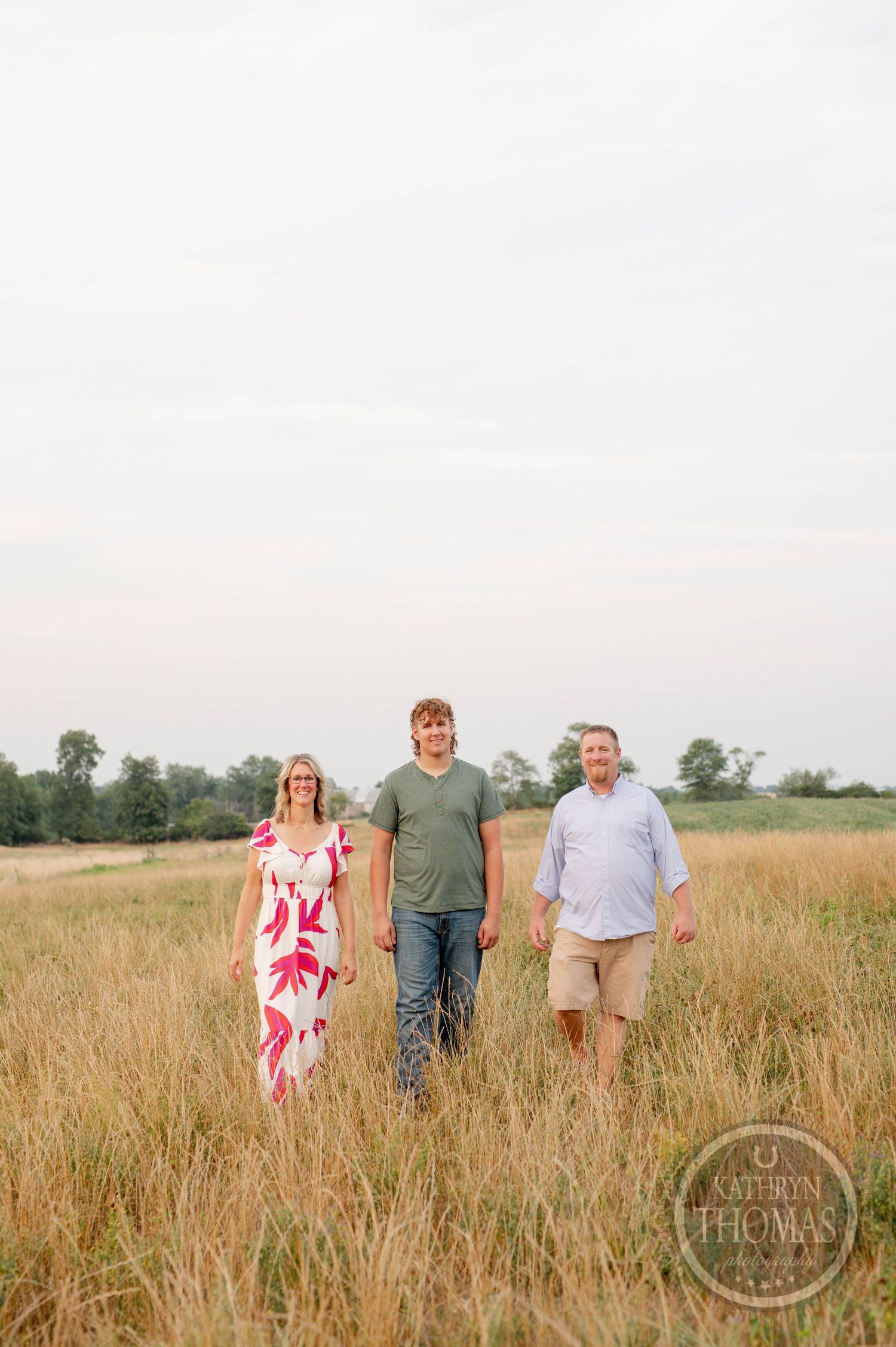 Three people standing in a field of tall grass during sunset with trees in background.