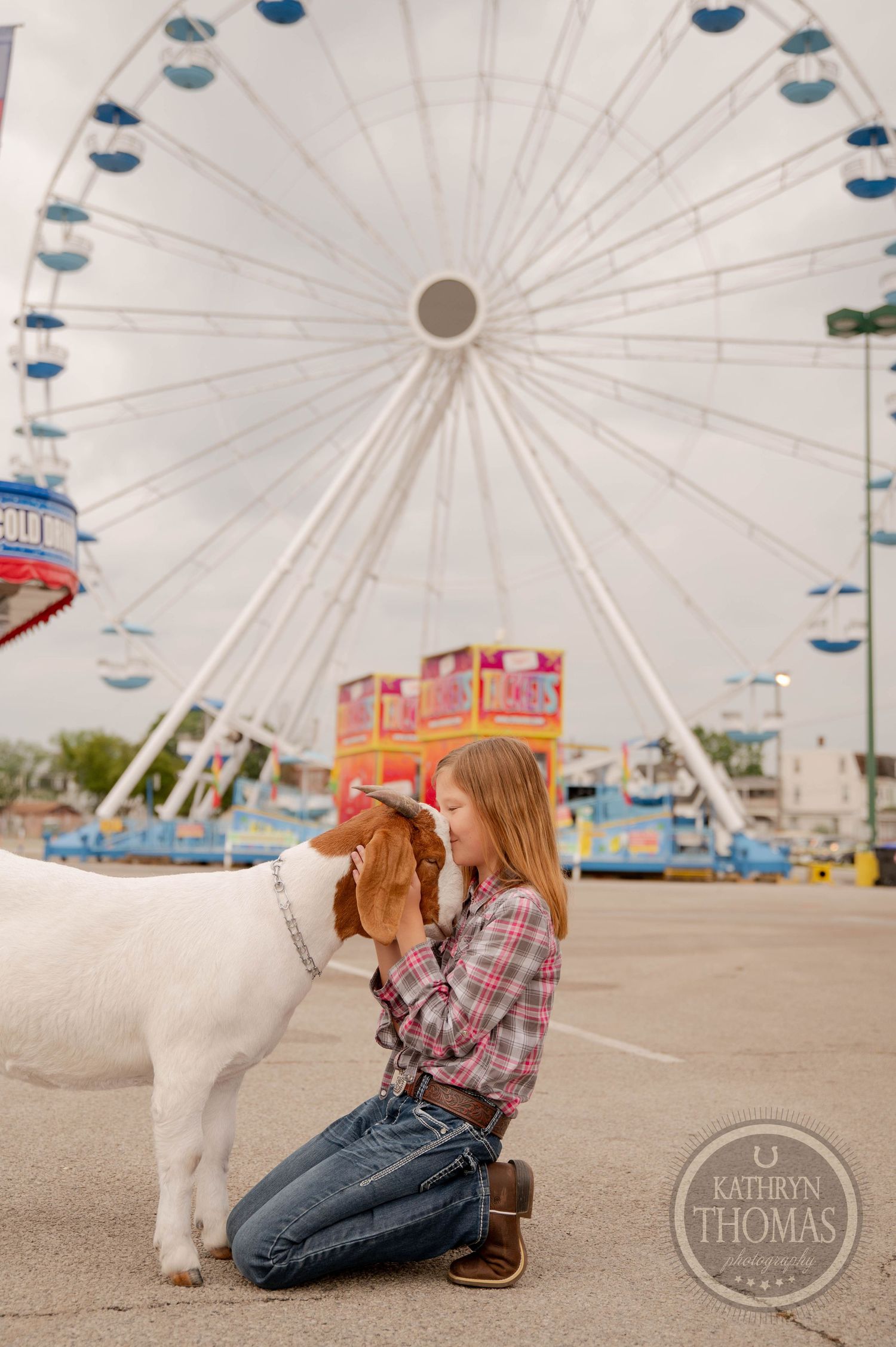A white goat poses with someone in front of a large ferris wheel at a carnival.