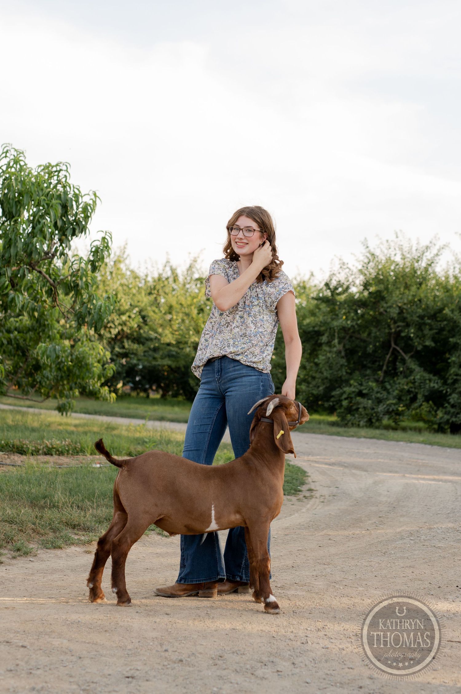 A brown goat stands attentively next to its handler on a country path with natural landscaping.
