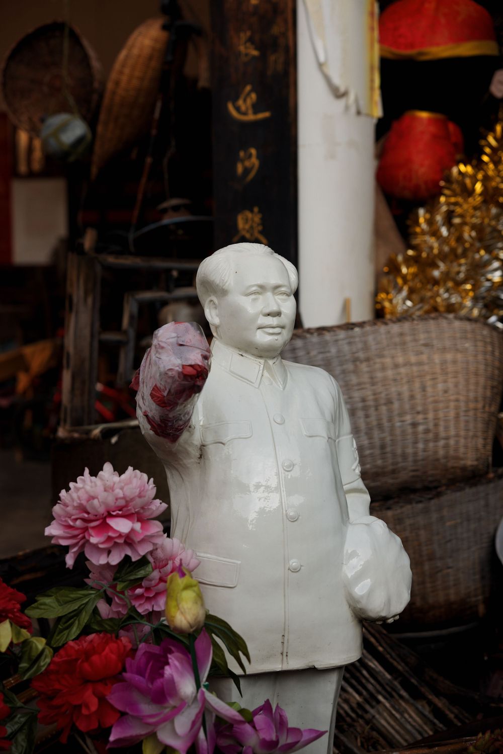 A white porcelain Mao statue with colorful flowers in front sits among wicker baskets and Asian decor.