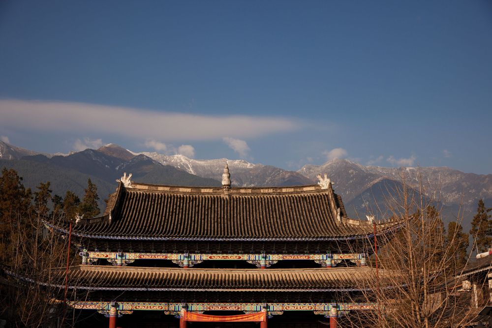 Traditional Chinese temple roof architecture against a backdrop of snow-capped mountains and blue sky.