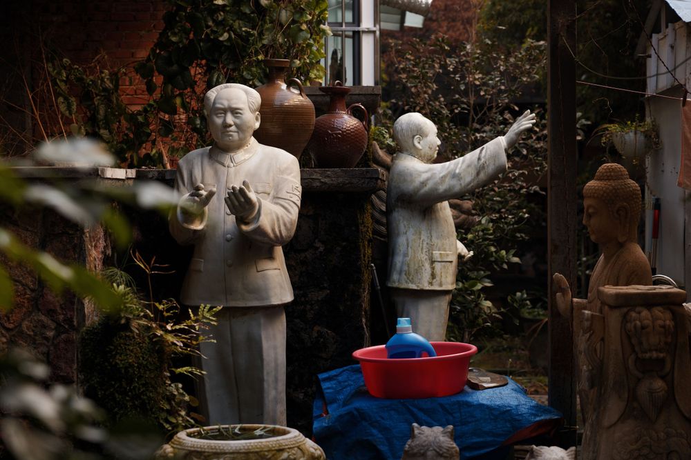 Weathered stone statues in garden setting with red and blue plastic containers in foreground.