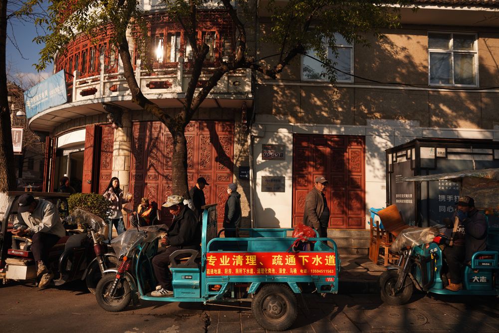 Street vendors with scooters and a red cart gather outside shops on a sunny day in an urban neighborhood.