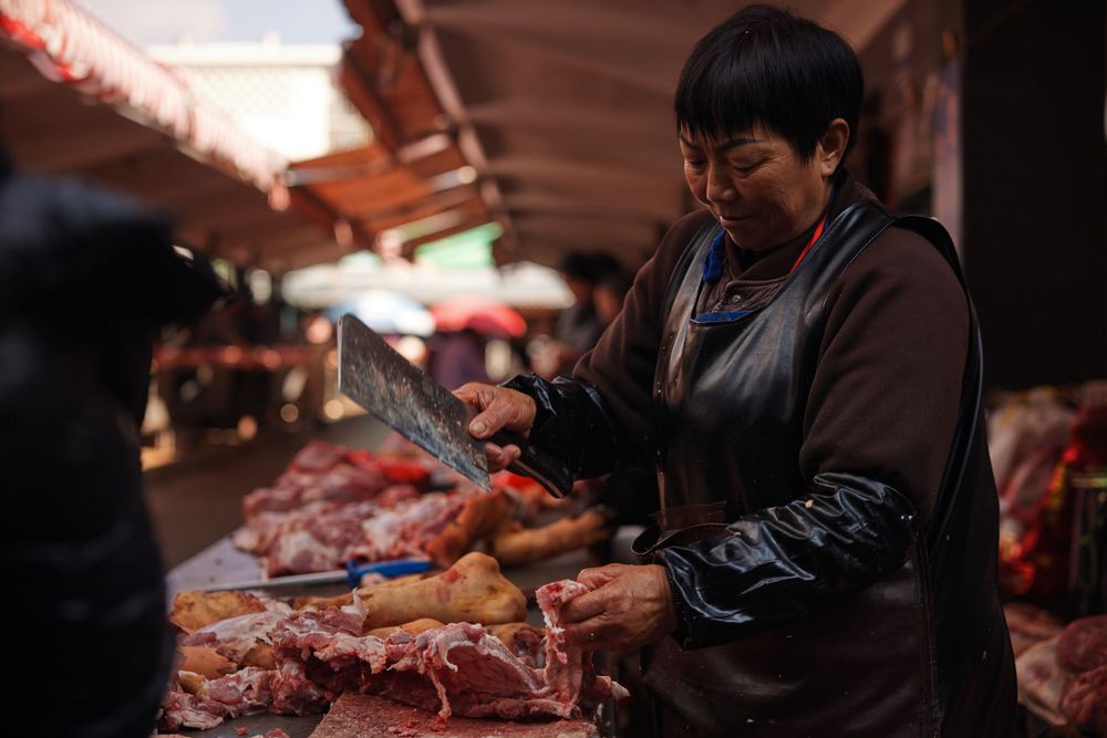 Butcher in dark apron slices raw meat at an indoor market stall under warm lighting.