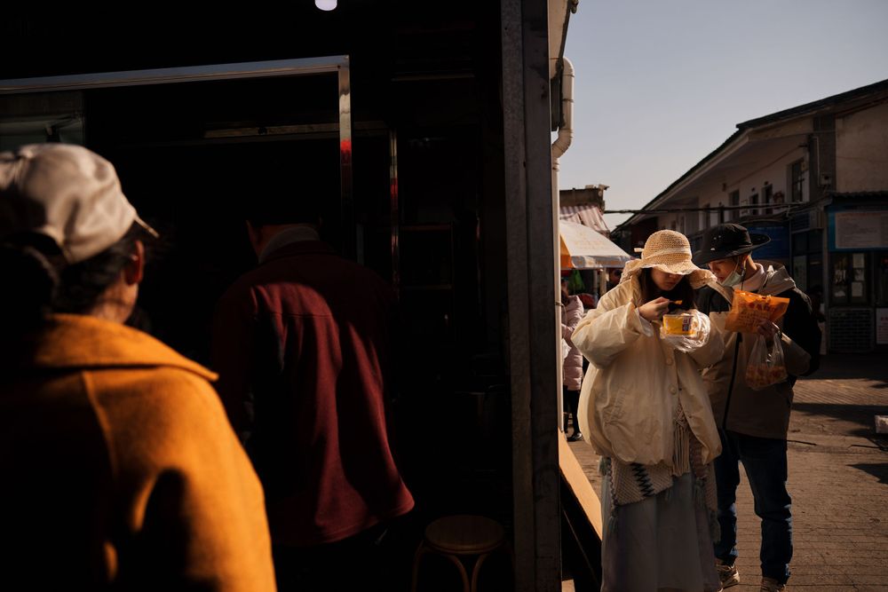 People waiting at a train station platform during sunset, casting long shadows in warm golden light.