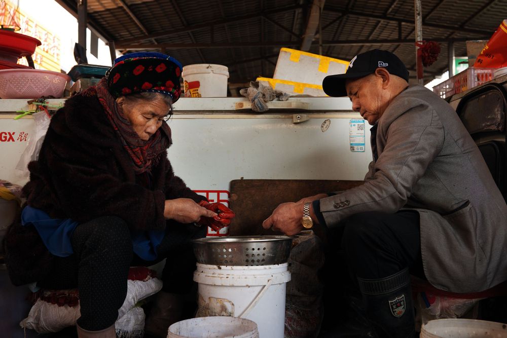 Two people sort through goods in plastic containers at a covered market stall in Asia.