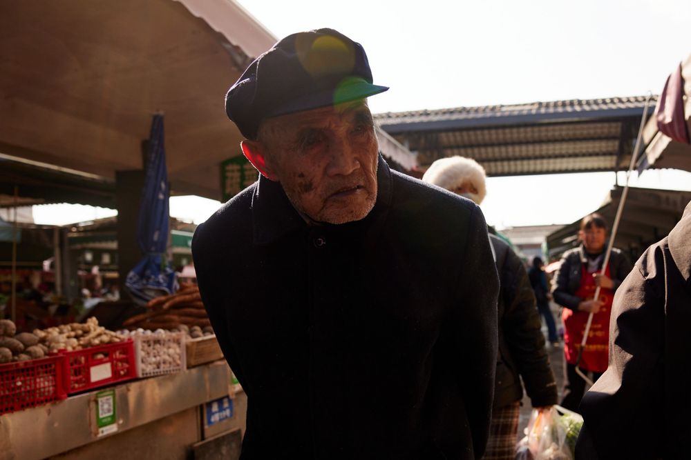 An elderly person wearing a dark cap browses items at a covered market or vendor stall.