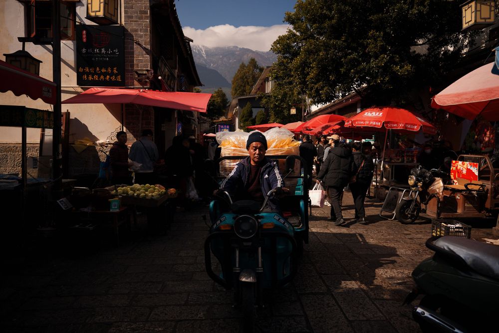 People relax at outdoor cafe tables under red umbrellas on a scenic mountain street in late afternoon light.