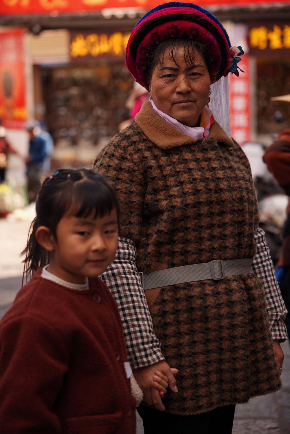 A parent and child in warm winter clothing stand together on a street with glowing red signs in the background.